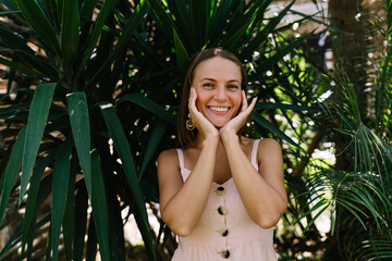 Half length portrait of cheerful female tourist dressed in casual wear smiling at camera during daytime leisure for exploring tropical environment with evergreen plants, happy woman on vacations