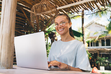 Portrait of happy graphic designer on classic eyeglasses using netbook computer during remote working outdoors, cheerful female copywriter with laptop enjoying digital nomad and freelance lifestyle