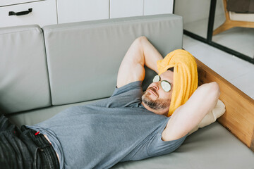 young handsome man is lying on sofa in living room and resting with towel on head and cucumber slices mask on eyes, male appearance care, relax at home, treat yourself