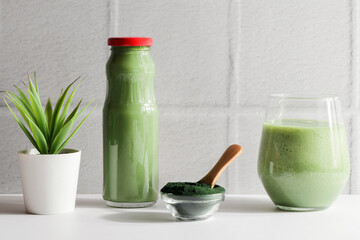 green smoothie in glass and spirulina algae powder in bowl with spoon,flower on white ceramic tile background.self care and healthy lifestyle concept.
