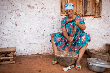 African woman cooks using a mortar and pestle, she is dressed in a traditional dress and she is in...