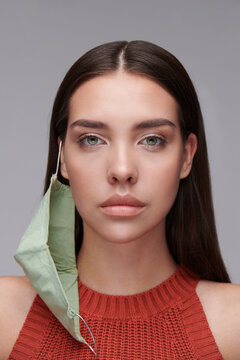 Portrait Of Serious Young Woman With Cloth Mask Hanging On Ear Against Gray Background, Coronavirus Concept