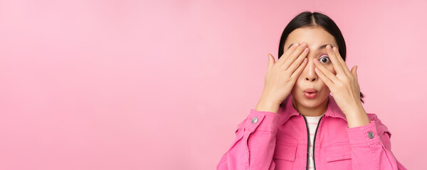 Fototapeta Close up portrait of young asian girl looking surprised, express amazement and wonder, peeking through fingers, standing over pink background obraz