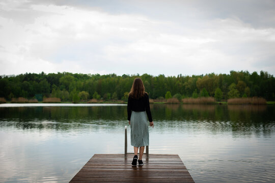 A young slender woman in cloudy weather walks to the edge of a wooden pier