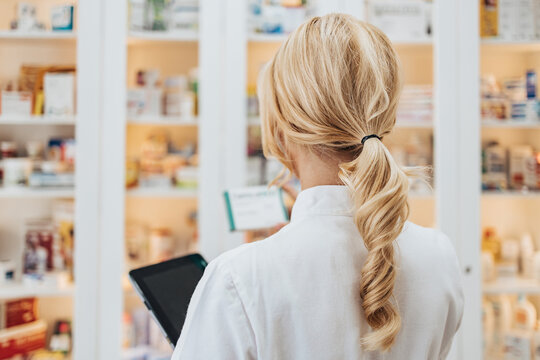 Young and attractive female pharmacist working in a drugstore. She is confident and serious. View from the back.