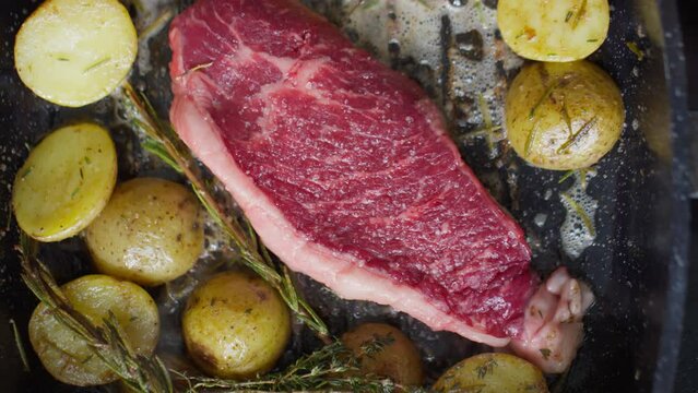 Close up top down shot of beef steak and potatoes being cooked on hot grill pan with butter and herbs