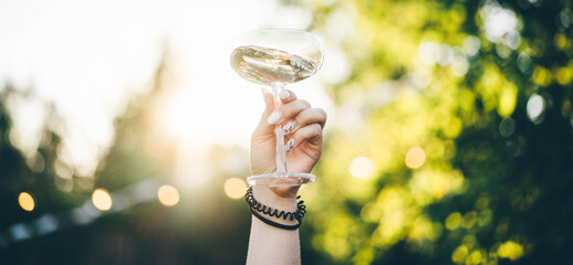 Woman hand holding a glass of white wine on the blurred background of green park.