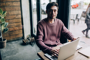 Portrait of Caucasian hipster guy in casual wear using modern laptop computer and digital cellphone...