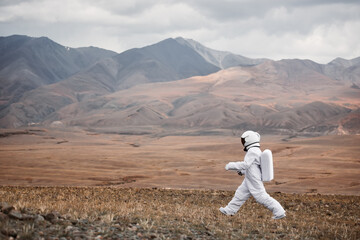 An astronaut explores an unknown red planet. Cloudy sky and mountains in the background.