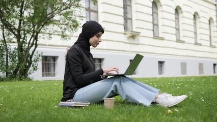 Elegant muslim arab islamic student girl in hijab. Woman freelancer user uses laptop sitting on green grass in park near university working online studying remotely chatting browsing shopping outdoor.
