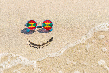 Fototapeta na wymiar Sunglasses with flag of Grenada State on a sandy beach. Nearby is a sea lightning and a painted smile. The concept of a successful vacation in the resorts of Grenada.