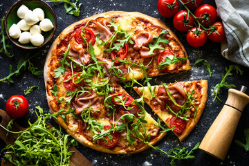 Pizza on black background. Traditional italian pizza with prosciutto, cheese, tomatoes and arugula with ingredients. Top view.