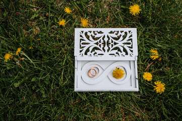 A beautiful white box with an infinity symbol with golden wedding rings lies on green grass with blooming yellow dandelions. Festive photo.