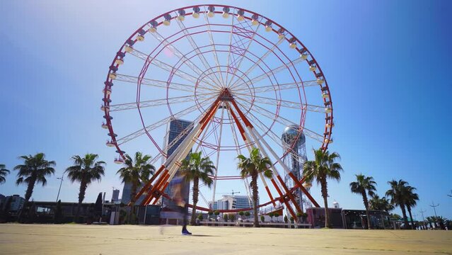Slow-motion Ferris wheel against blue sky. Batumi. Georgia. Amusement park on hot summer day. TIMELAPSE OF THE FERRIS WHEEL, WHICH ROTATES IN ACCELERATED MOTION. Palm trees, a resort town.