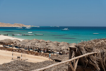 Holidays in Egypt. Beach with umbrellas made of dry palm leaves, desert and yachts in the blue water of the Red Sea. Vacation and Holidays in Egypt.