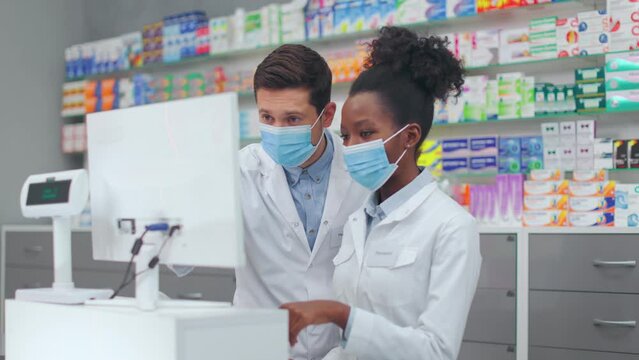 Two Drugstore Workers In Face Masks Having Talk While Standing Together Behind Counter. Caucasian Male Pharmacist Teaching African American Female Assistant To Work On Computer In Pharmacy.