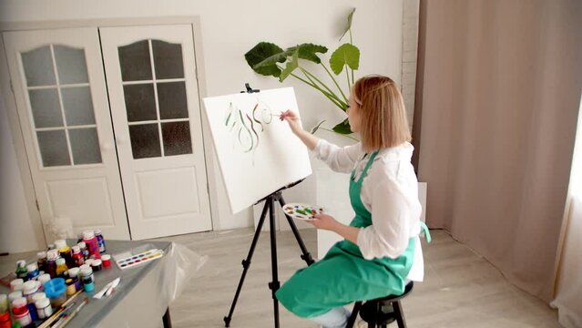 Woman in an apron drawers with brush on canvas, there are jars of paint and different brushes at table, side view. Woman draws in living room on an easel. Hobby concept