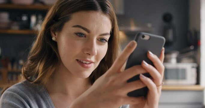 a young woman using a phone at home. A young woman sitting in her kitchen browsing online apps on her cellphone, smiling. A young woman relaxing at home sending a text message on her mobile phone