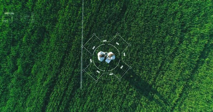 Top view of male and female scientists in white lab coats standing among green field with digital tablet and documents in hands. Two scientists activating virtual screen with holograms over crops.
