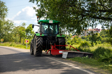 Big modern industrial tractor machine cutting green grass with mowing equipment along country...