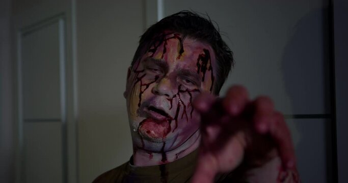 Scary zombie man with blood stains and streaks on face stretches hand to camera. Yellow and blue colors on face of zombie with blood traces closeup
