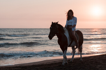 Woman in summer clothes enjoys riding a horse on a beautiful sandy beach at sunset. Selective focus 