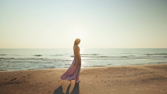 Panning: Pretty woman in long dress walking on wet sand against sunset sea background