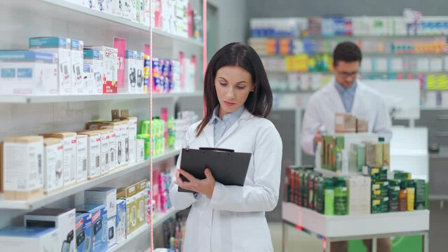 Young female pharmacist writing notes while working in modern drugstore. Сaucasian woman checking products reserve on pharmacy shelves using clipboard.