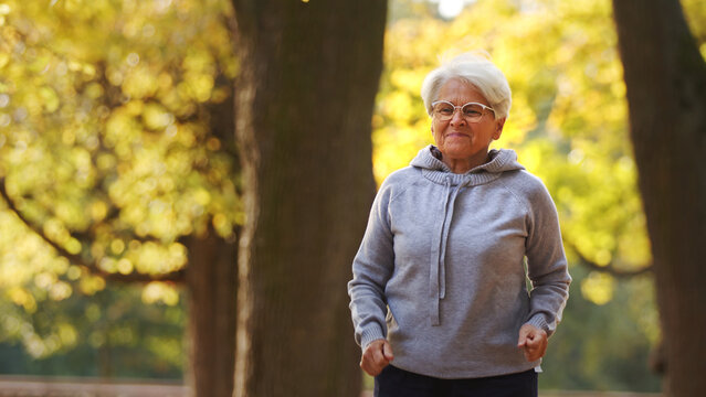 Jogging in park. Retired gray-haired caucasian lady in gray hoodie runs around her favorite park. Wellbeing concept. High quality photo