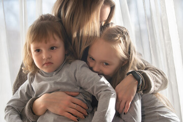 Portrait of a beautiful young woman with two little daughters at home against the background of the window. Family spend leisure time in cozy home environment