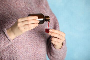 Woman pouring cough syrup into measuring cup on light blue background, closeup