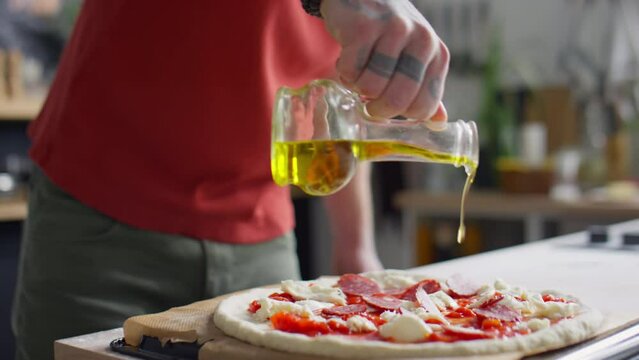 Cropped shot of male food blogger pouring olive oil over pizza before baking and showing devil horn hand gesture on camera while cooking in kitchen