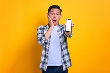 Shocked young Asian man in plaid shirt showing mobile phone blank screen recommending app isolated on yellow background