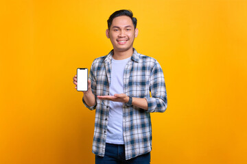 Smiling young Asian man in plaid shirt showing mobile phone blank screen recommending app isolated on yellow background