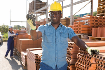 African-american builder standing beside brick stacks in outdoor construction material warehouse.