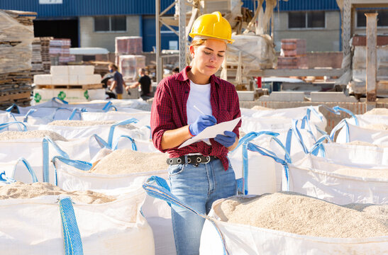 Young Female Worker Standing Amongst Bags With Bulk Construction Materials In Outdoor Warehouse. She's Holding Paper And Pen In Hands.