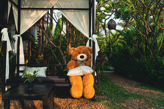 Empty dining table with teddy bear sitting on a long chair in the green garden area, decoration design with natural background in summer, feeling relax with flower and plant.