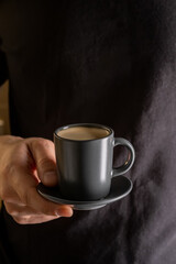 A man's hand holds a cup of hot espresso coffee against the background of a black T-shirt.