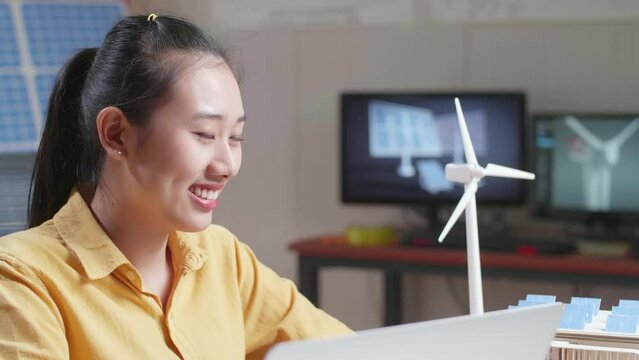 Asian Woman Smile To Camera While Working With A Laptop Next To The Model Of A Small House With Solar Panel
