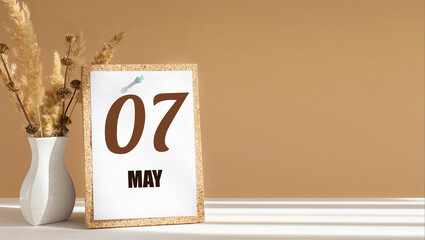 may 7. 7th day of month, calendar date.White vase with dead wood next to cork board with numbers. White-beige background with striped shadow. Concept of day of year, time planner, spring month