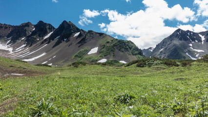 Fototapeta premium A picturesque mountain range against a background of blue sky and clouds. Snowmelt is visible on the slopes. Juicy green grass grows in the meadow. Kamchatka. Vachkazhets
