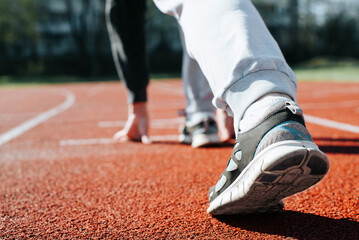 Close-up of woman practicing running in stadium, standing at low start on sunny day. Sportswoman exercising outdoors. Back view, selective focus on sneaker