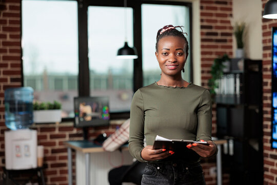 Production department team leader standing in digital art studio. Confident creative art designer standing in office workspace having clipboard paperwork documentation while smiling at camera.