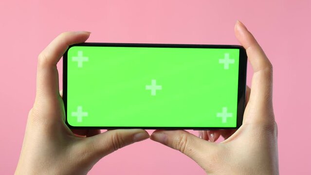 close-up of female hands holding smartphone with blank green screen showing copy space for app or website design, on pink background , mockup.