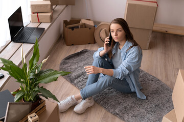 Young woman talking on phone sitting in new apartment moving in, student sitting on the floor with boxes, relocation to a new home