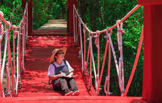 An Asian female forester taking notes while sitting on red suspension bridge in mangrove forest at natural parkland