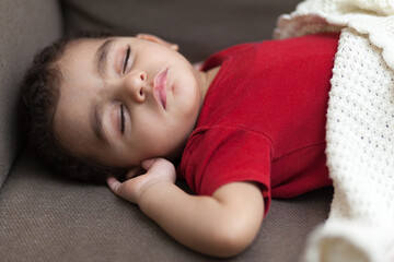 A little boy in a red overalls sleeps on a couch, covered with a white blanket. High quality photo