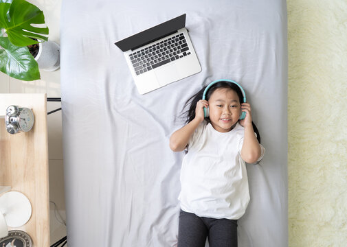 Top view Asian child little girl wearing headphones with laptop computer on the bed