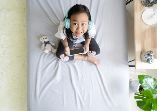 Top view Asian child little girl play smartphone with wearing headphones on the bed
