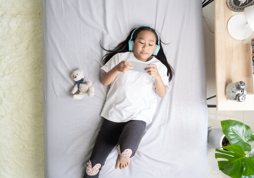 Top view Asian child little girl play smartphone with wearing headphones on the bed